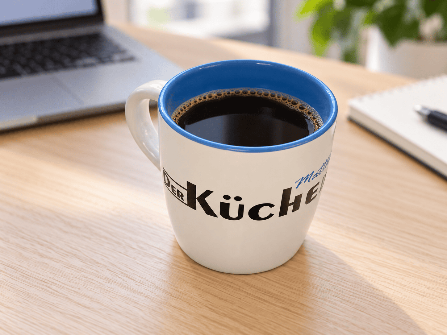Weiße Logotasse mit blauem Innenraum und schwarzem Kaffee auf einem Schreibtisch neben einem Laptop